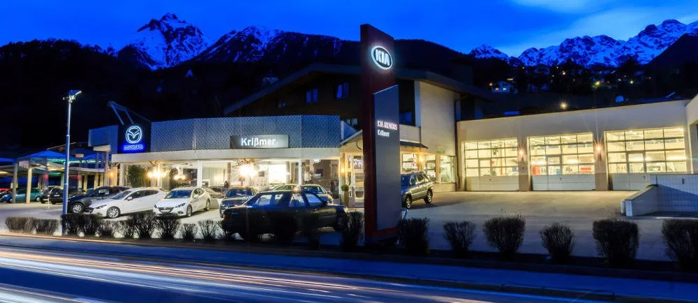 Das beleuchtete Autohaus in der Dämmerung, mit Mazda-Säule und KIA-Säule und den hellen Werkstatttoren