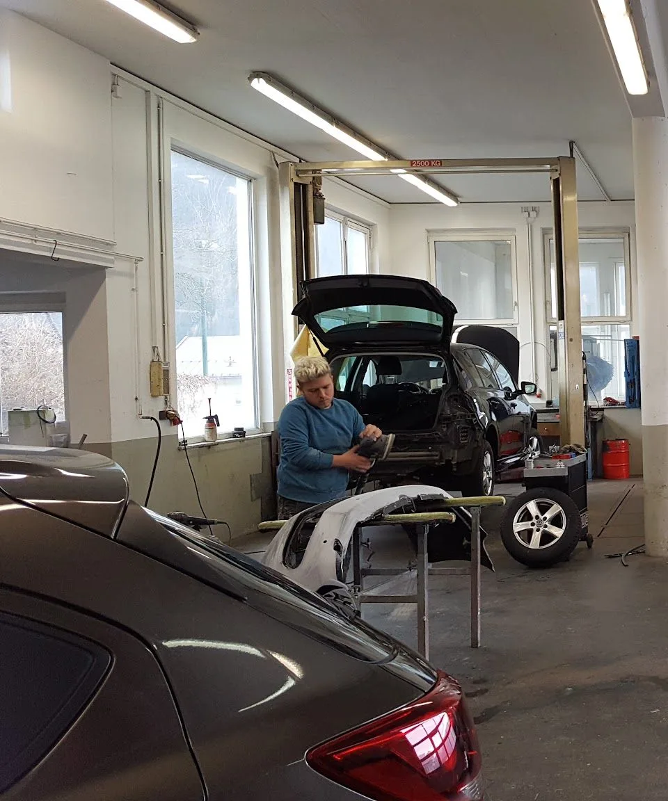 Ein Mitarbeiter schleift in der Werkstatt ein Karosserieteil, dahinter steht ein Fahrzeug auf der Hebebühne