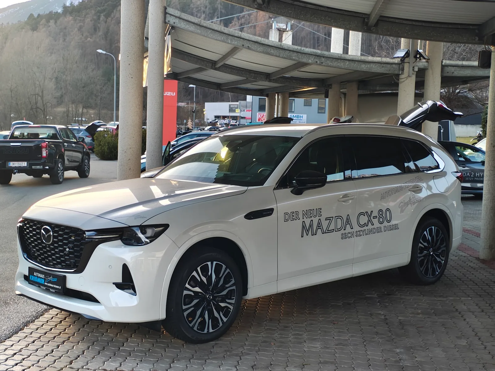 Ein weißer Mazda CX-80 als beschrifteter Vorführwagen unter dem Vordach des Autohauses
